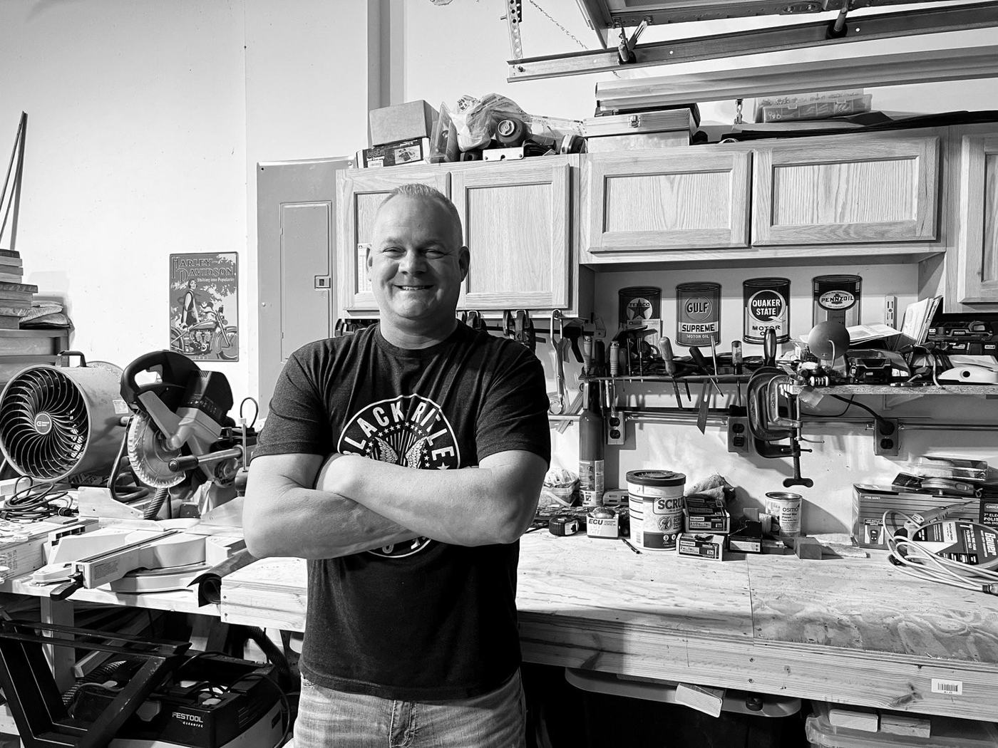 Dan Goodman standing with arms crossed at his workbench, black and white